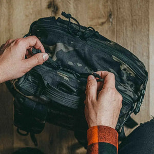 Hands opening a camouflage 12 Hour Plus Backpack with a zipper.