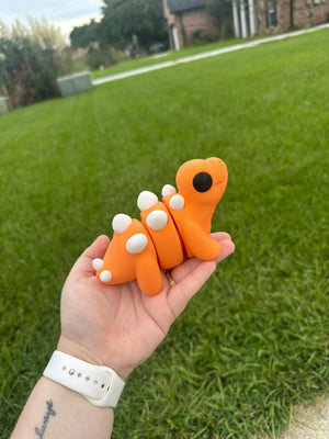 Orange 3D Printed Stegosaurus Fidget Toy on Hand with Green Grass Background - Main Street Collective
