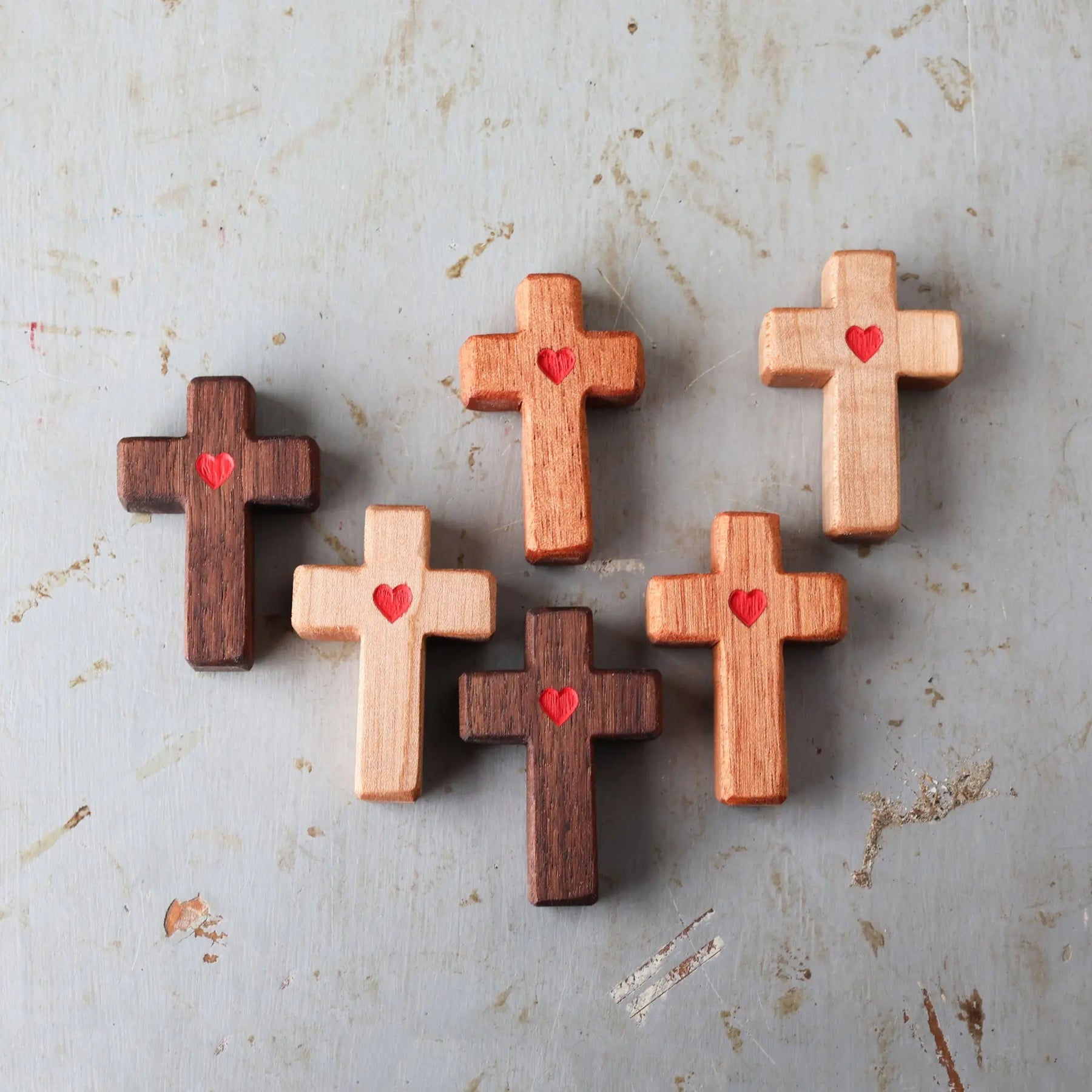 Wooden pocket crosses with carved red hearts arranged on a textured surface.