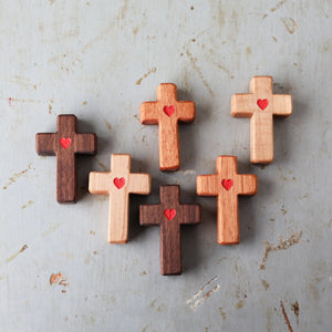 Wooden pocket crosses with carved red hearts arranged on a textured surface.