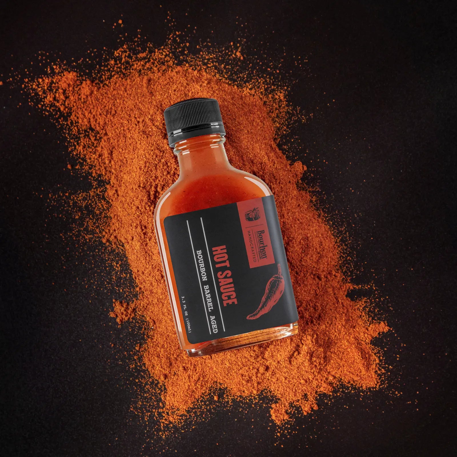Bourbon Barrel Aged Hot Sauce bottle surrounded by spilled chili powder on black surface.
