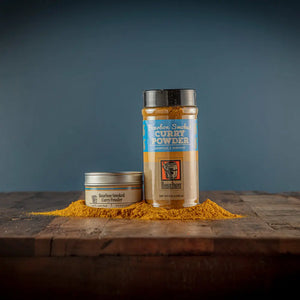 Two containers of Bourbon Smoked Curry Powder on a wooden surface with spilled powder.