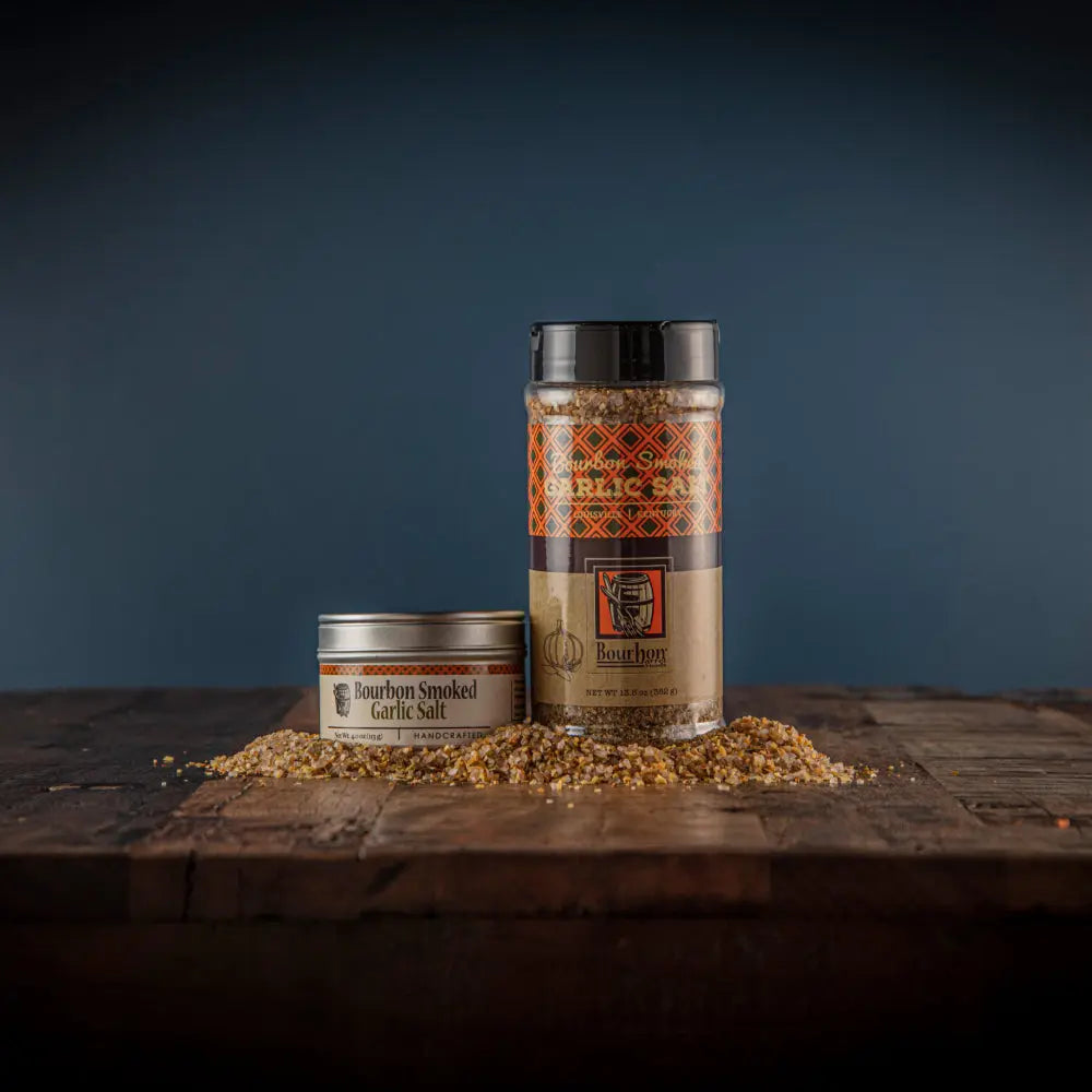 Bourbon Smoked Garlic Salt in tin and shaker containers on a wooden surface with scattered seasoning.
