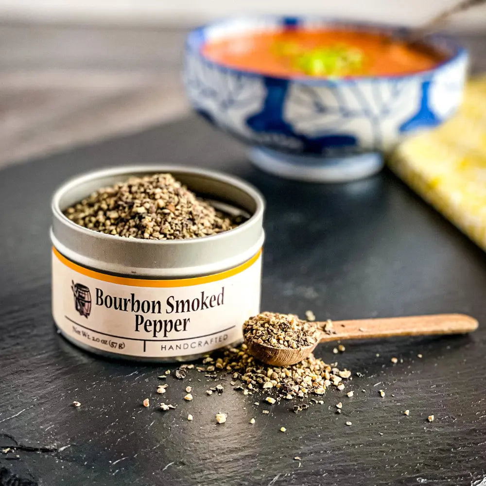 Bourbon Smoked Pepper in a tin with a wooden spoon on a dark surface