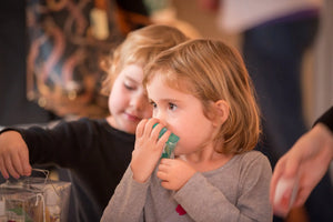 Young child holding green-wrapped soap slice near face, with another child behind