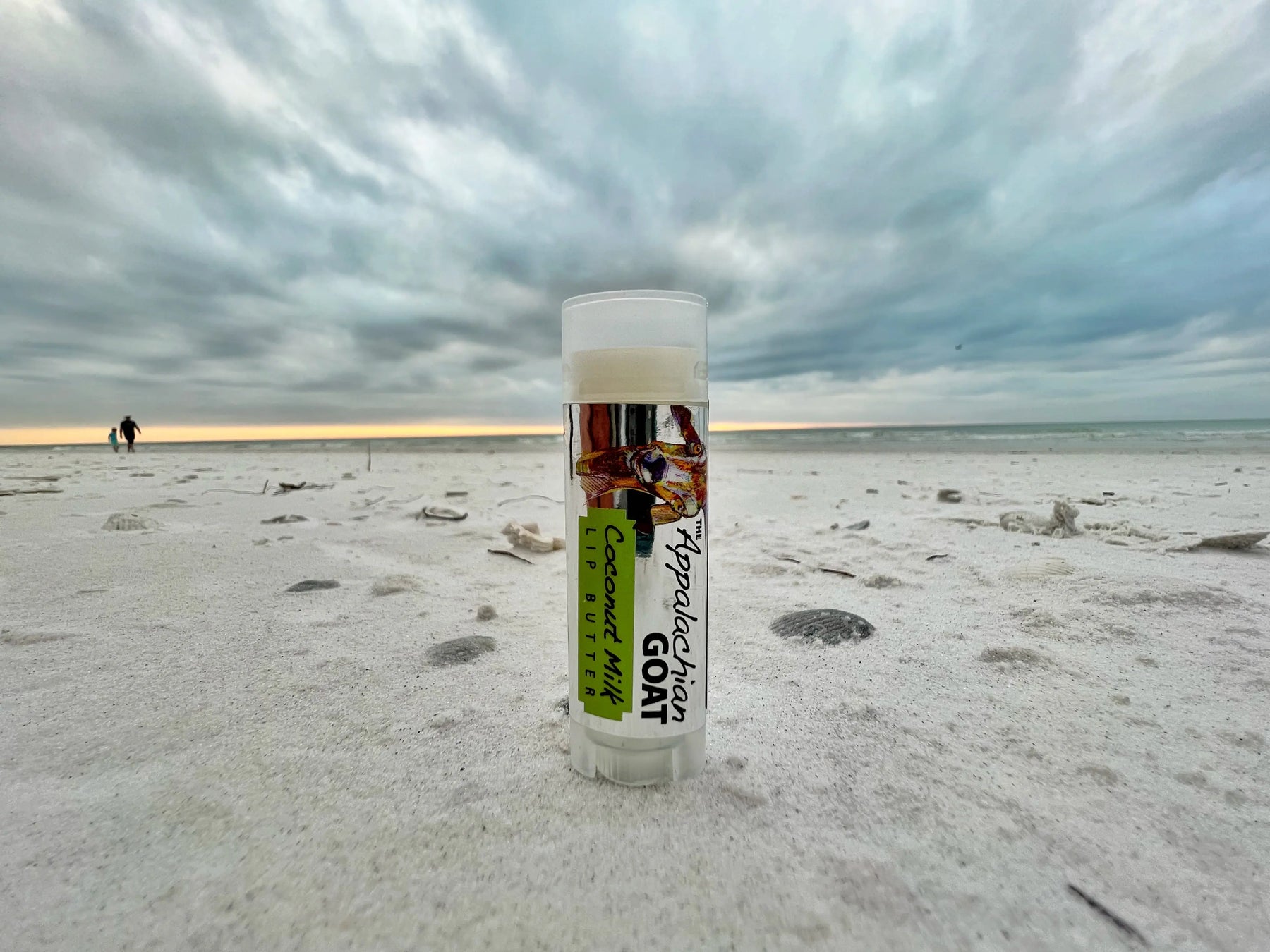 Appalachian Goat Coconut Milk Lip Butter tube on a sandy beach with cloudy sky.