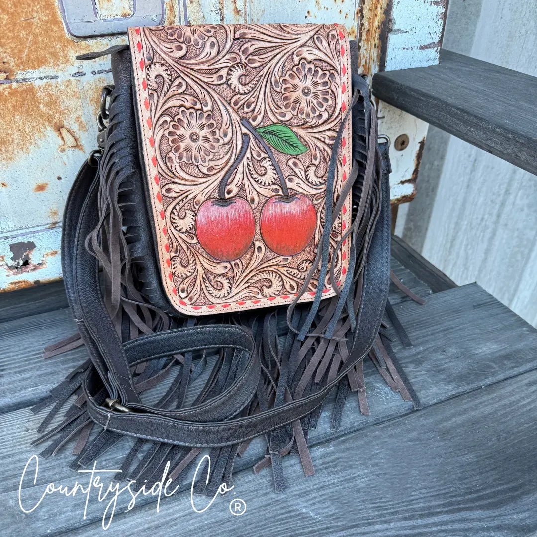 Brown leather crossbody purse with hand-tooled floral pattern and red cherries, featuring fringe detail.