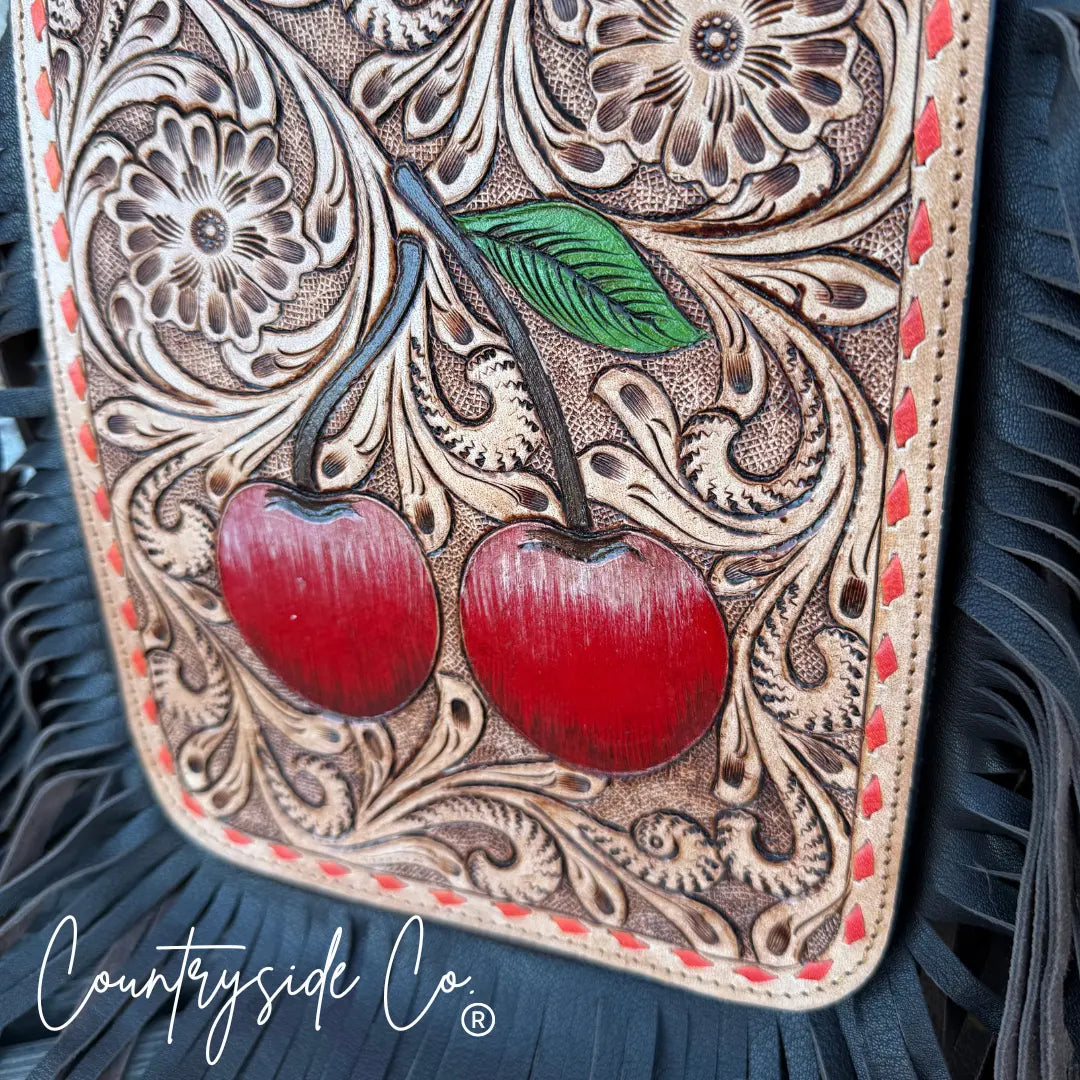 Hand-tooled leather purse with red cherries and floral patterns, featuring black fringe.
