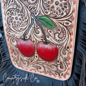 Hand-tooled leather purse with red cherries and floral patterns, featuring black fringe.