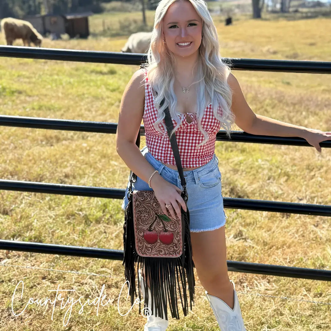 Woman wearing a red gingham top and denim shorts holding a tooled leather purse with cherries.