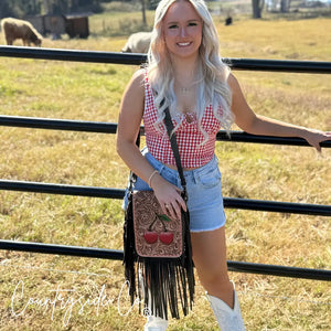 Woman wearing a red gingham top and denim shorts holding a tooled leather purse with cherries.
