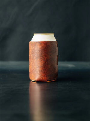 Leather coozie holding a white can with gold rim, sitting on a dark surface with a blurred dark background.  