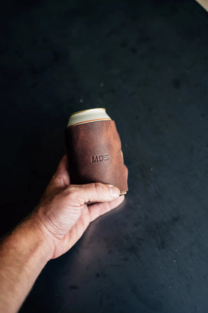 Hand holding a brown leather coozie with a can, "MDS" embossed, cognac nemesis, from Main Street Collective.