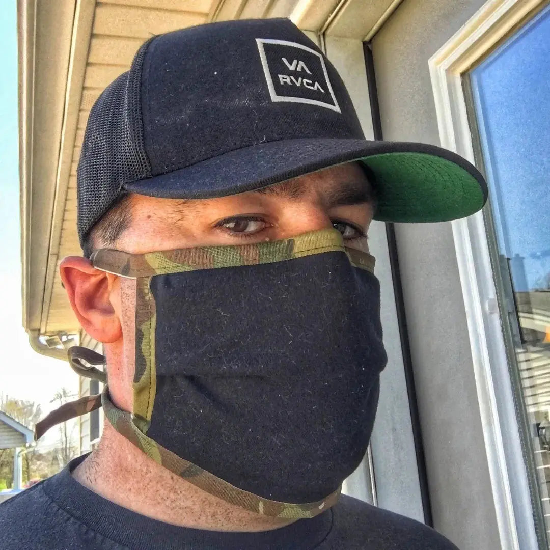 Man wearing a black cotton face mask with camouflage trim and a black baseball cap.