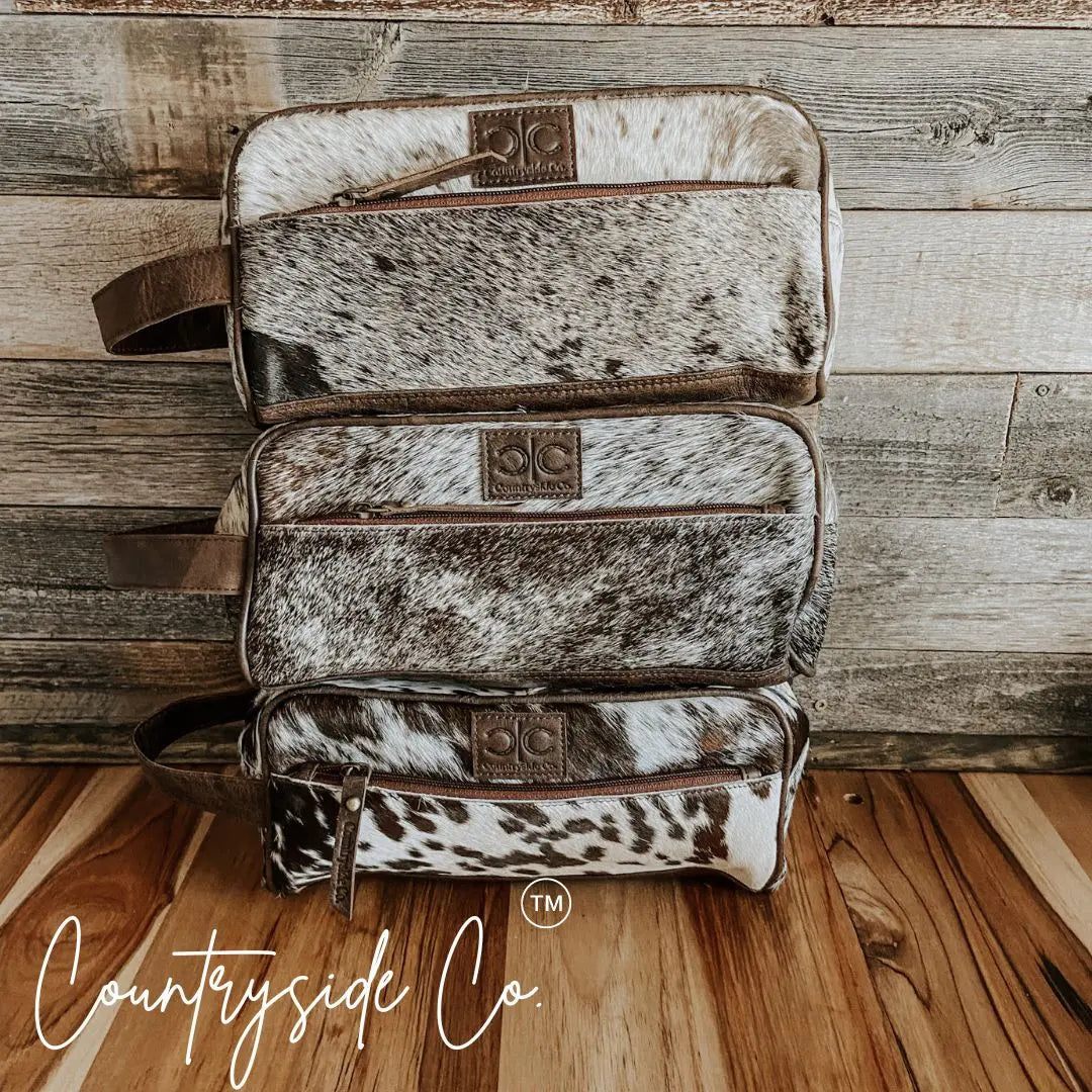 Stack of three Countryside Co. cowhide toiletry bags with leather trim.