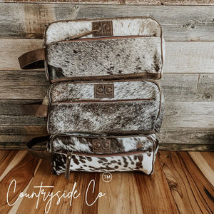 Stack of three Countryside Co. cowhide toiletry bags with leather trim.