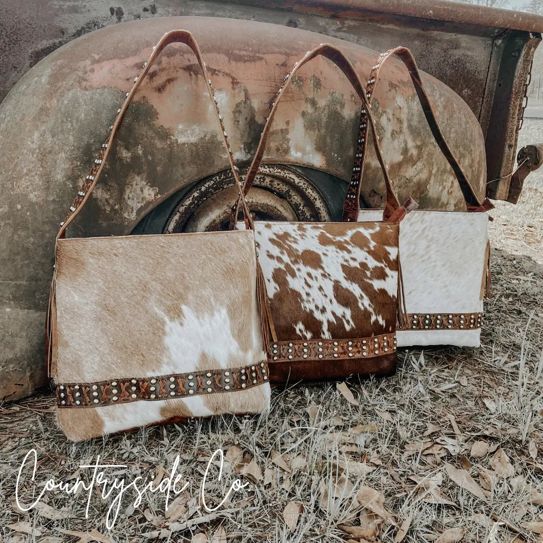 Three cowhide leather purses with braided straps and brass accents in various cow print patterns.