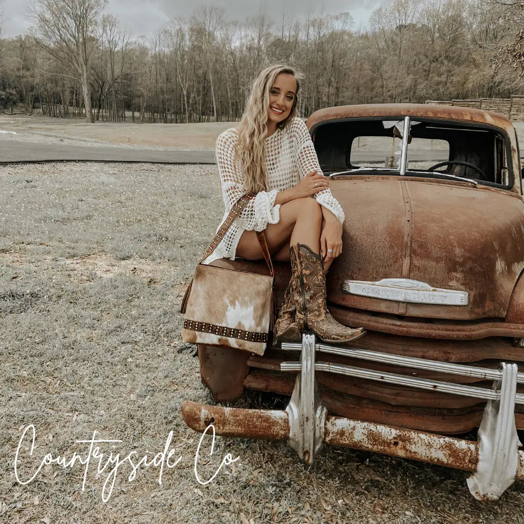 Woman in white dress and cowboy boots sitting on a rusty truck with a cowhide purse.