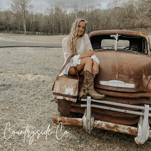 Woman in white dress and cowboy boots sitting on a rusty truck with a cowhide purse.