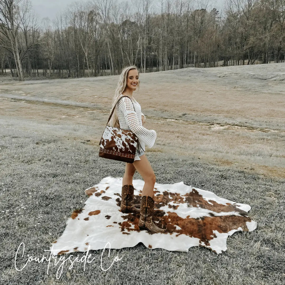 Woman with blonde hair wearing white crochet top and cowgirl boots holding a cowhide print leather purse outdoors.