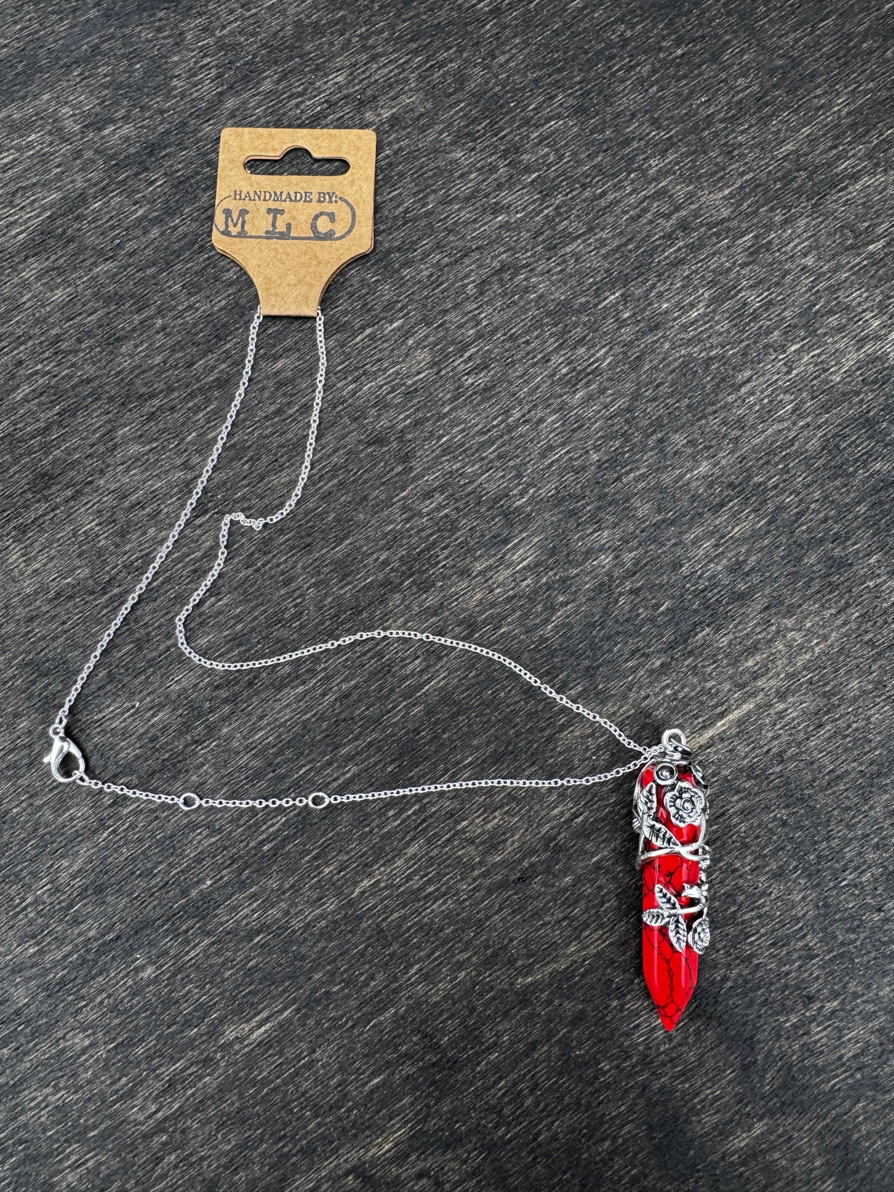 Red crystal pendant necklace with silver floral vines and a silver chain with Handmade By MLC tag on a dark wood background.
