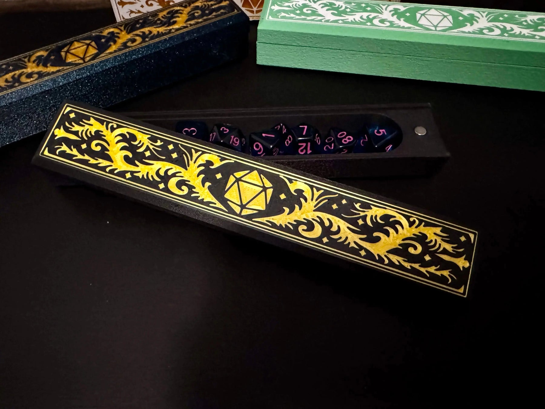 Open black 3D printed dice box with gold design, holding blue dice with pink numbers; other boxes visible in background.