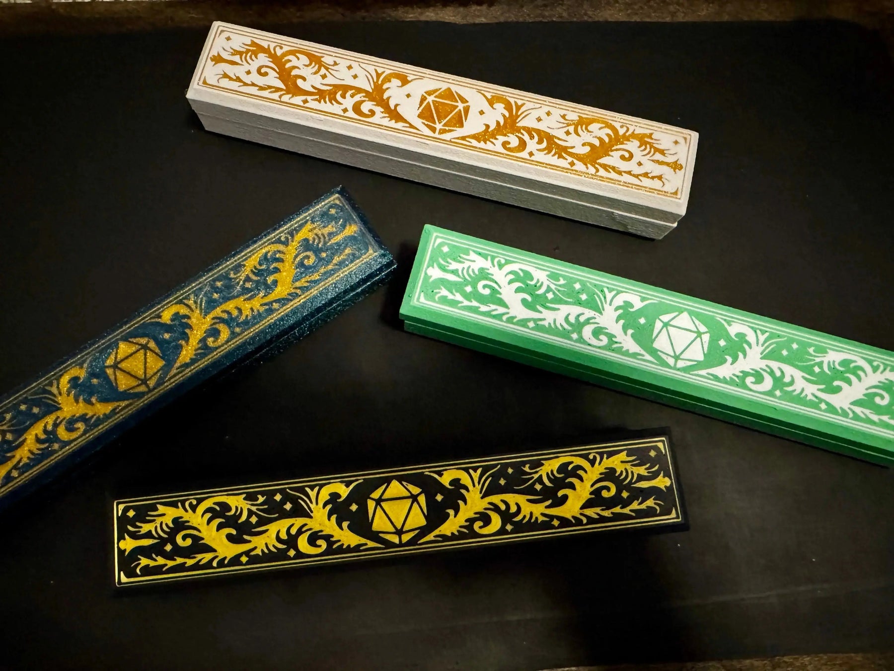 Four 3D-printed Adventurer dice boxes with different color schemes and a d20 symbol on a black surface.