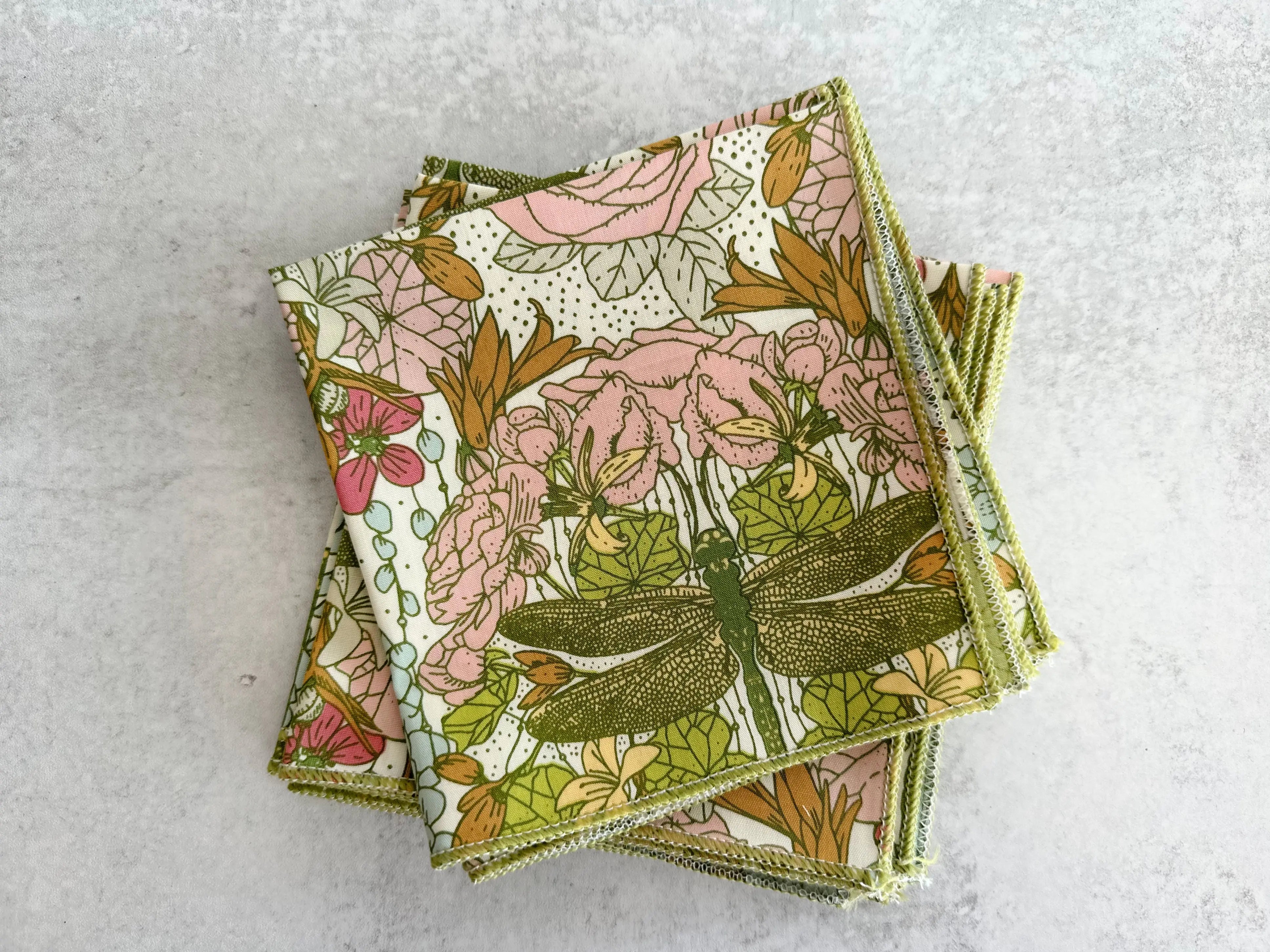 Stack of four cloth napkins with dragonfly and floral pattern in green, pink, and gold.