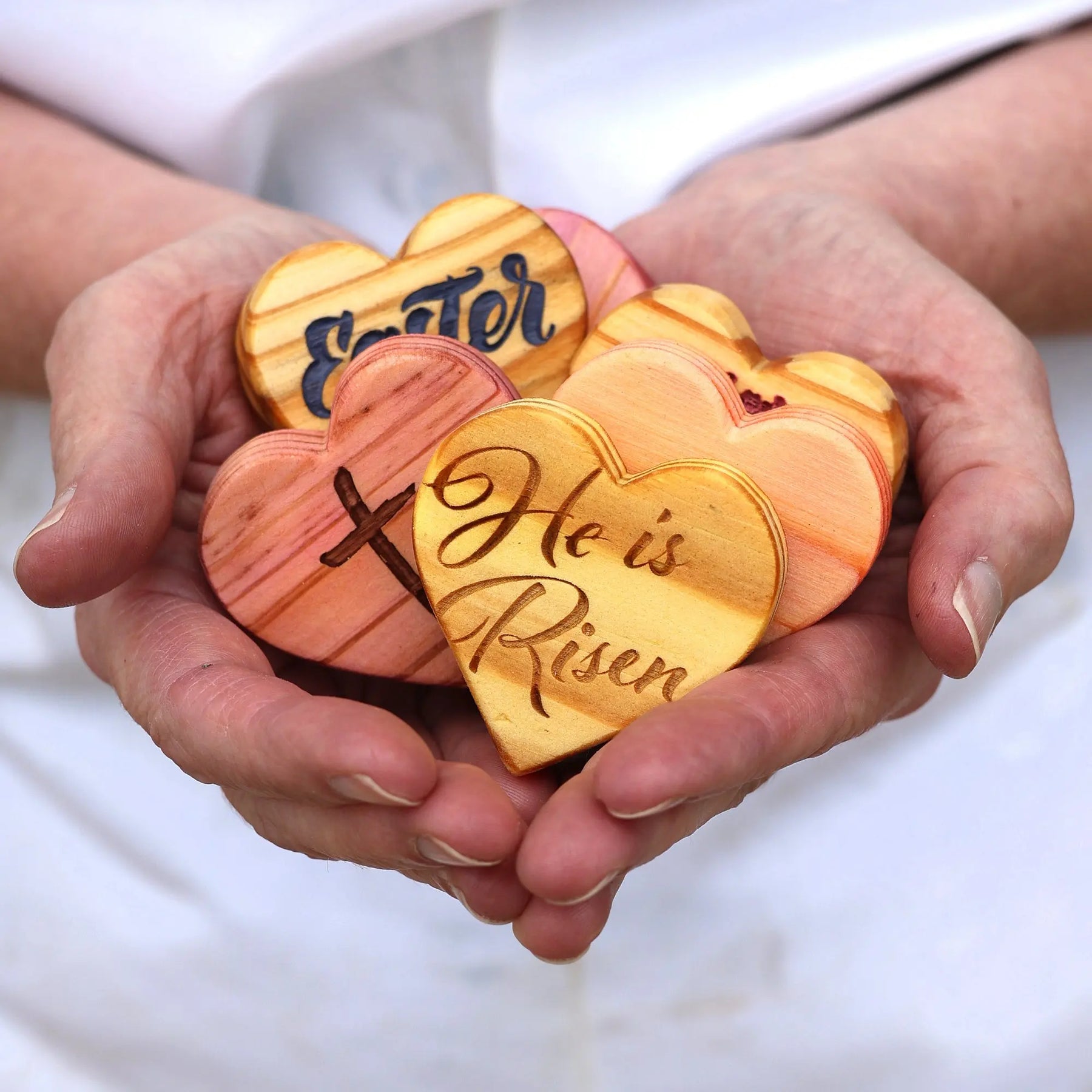 Engraved wooden hearts with "Easter" and "He is Risen" phrases held in hands.