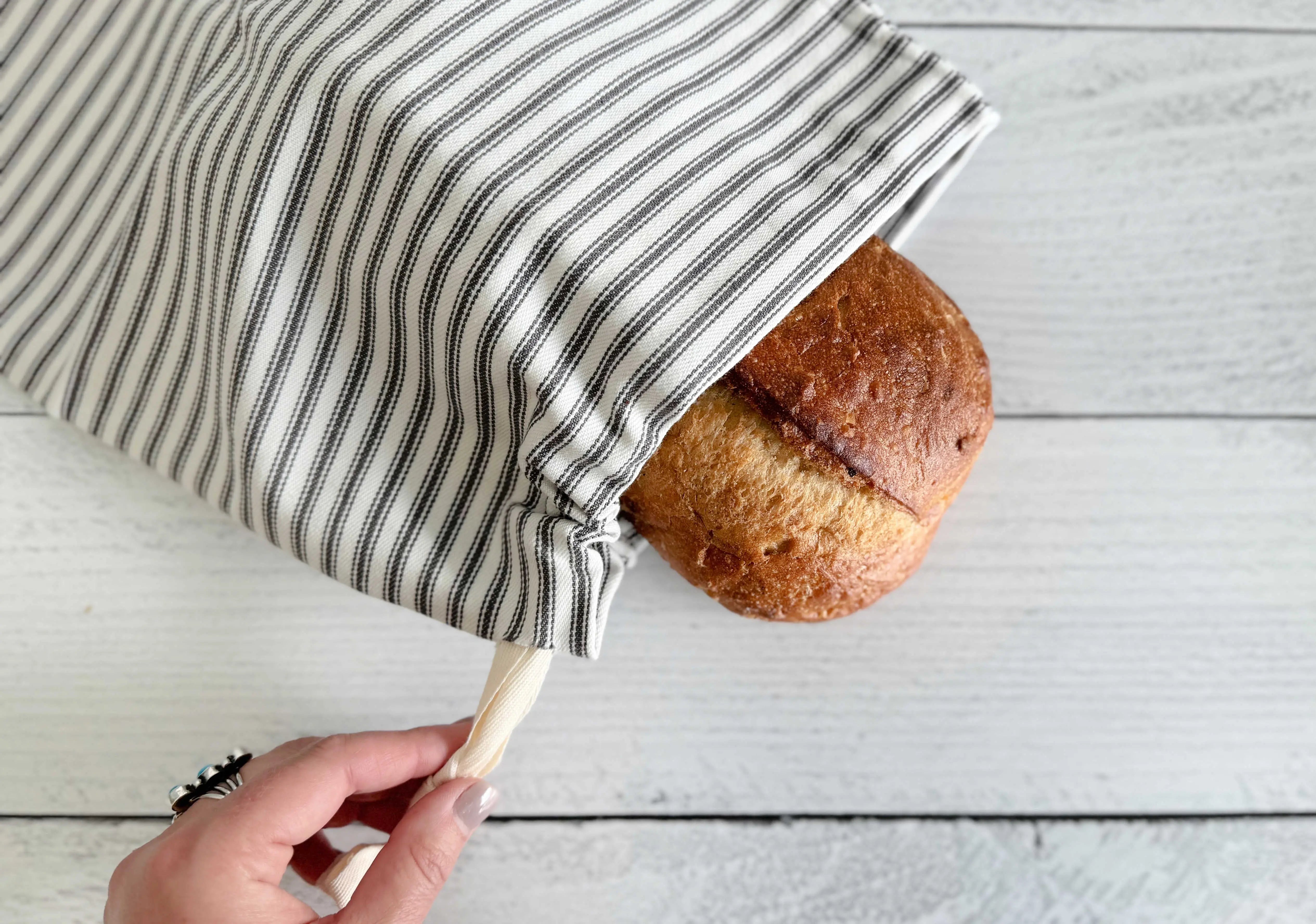 Grey ticking stripe bread bag with drawstring holding a loaf of bread.