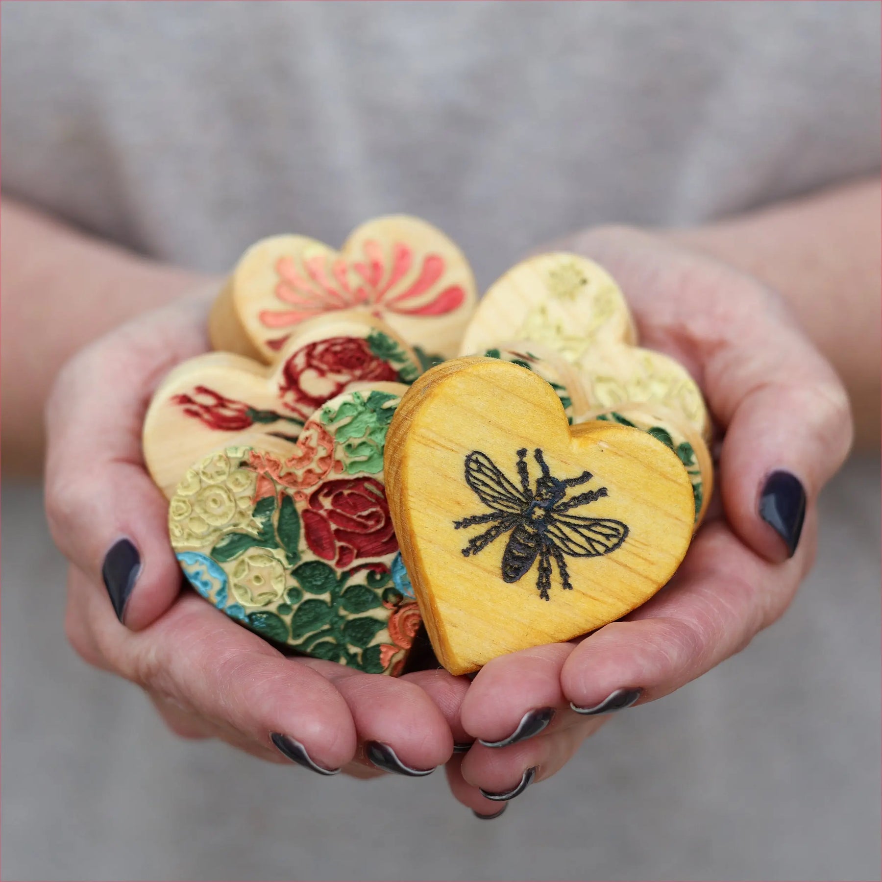 Hands holding heart-shaped wooden items with floral and bee designs.