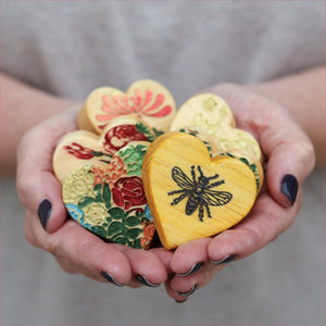 Hands holding heart-shaped wooden items with floral and bee designs.
