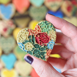 Hand holding a colorful heart-shaped item with raised floral patterns.