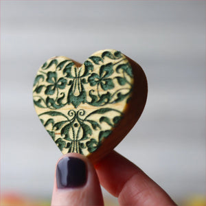 Engraved wooden heart with a dark green floral pattern held by fingers.