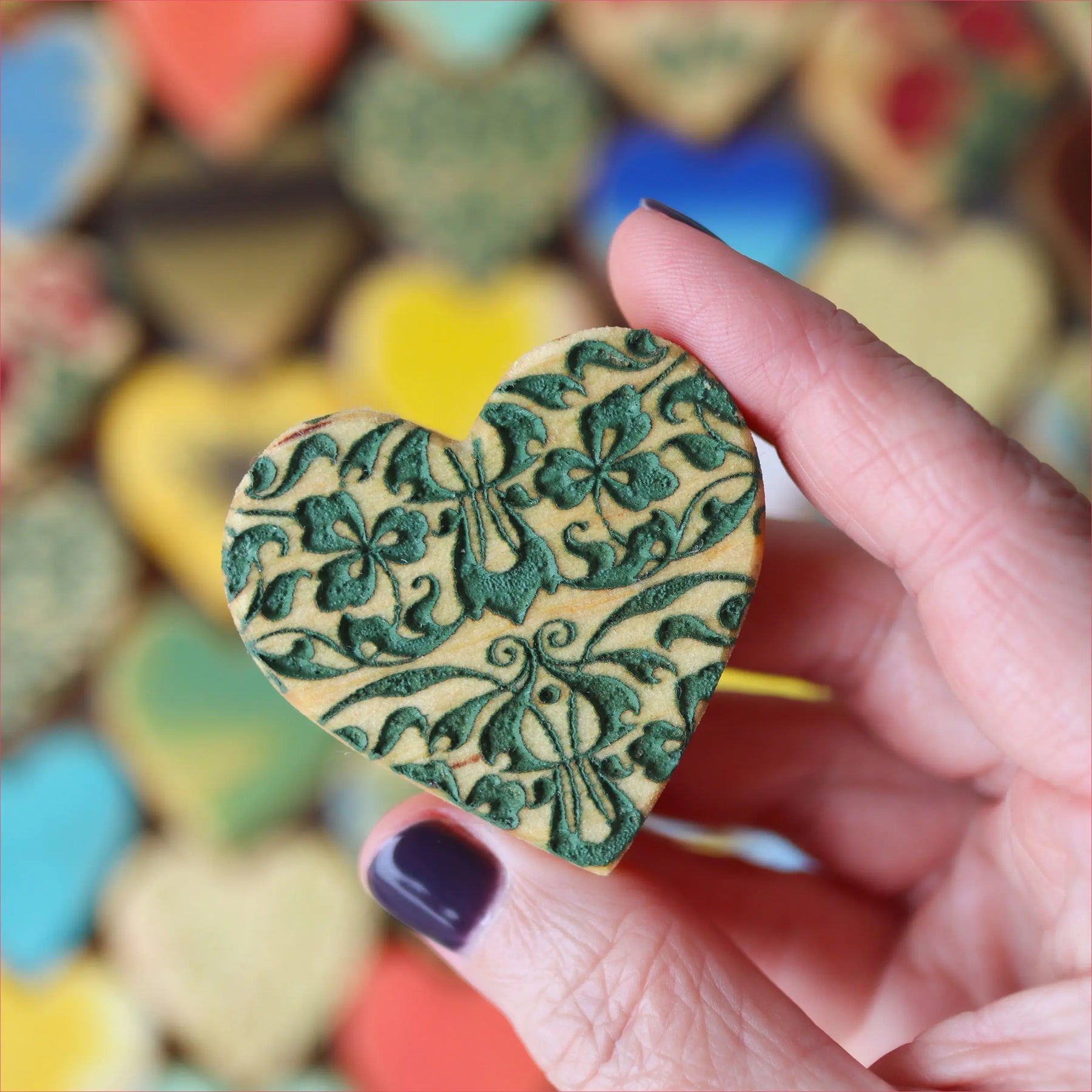 Hand holding a green engraved wooden heart with a floral pattern.