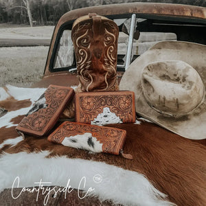 Brown tooled leather and cowhide wallet with floral tooling, displayed with a cowboy boot and hat.