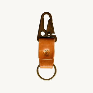 A key clip with brown leather strap, a brass lever clip, and an antique brass key ring on a white background.