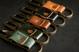 Keychains with leather straps (natural, black, brown, green) and brass clips/rings on a dark surface.
