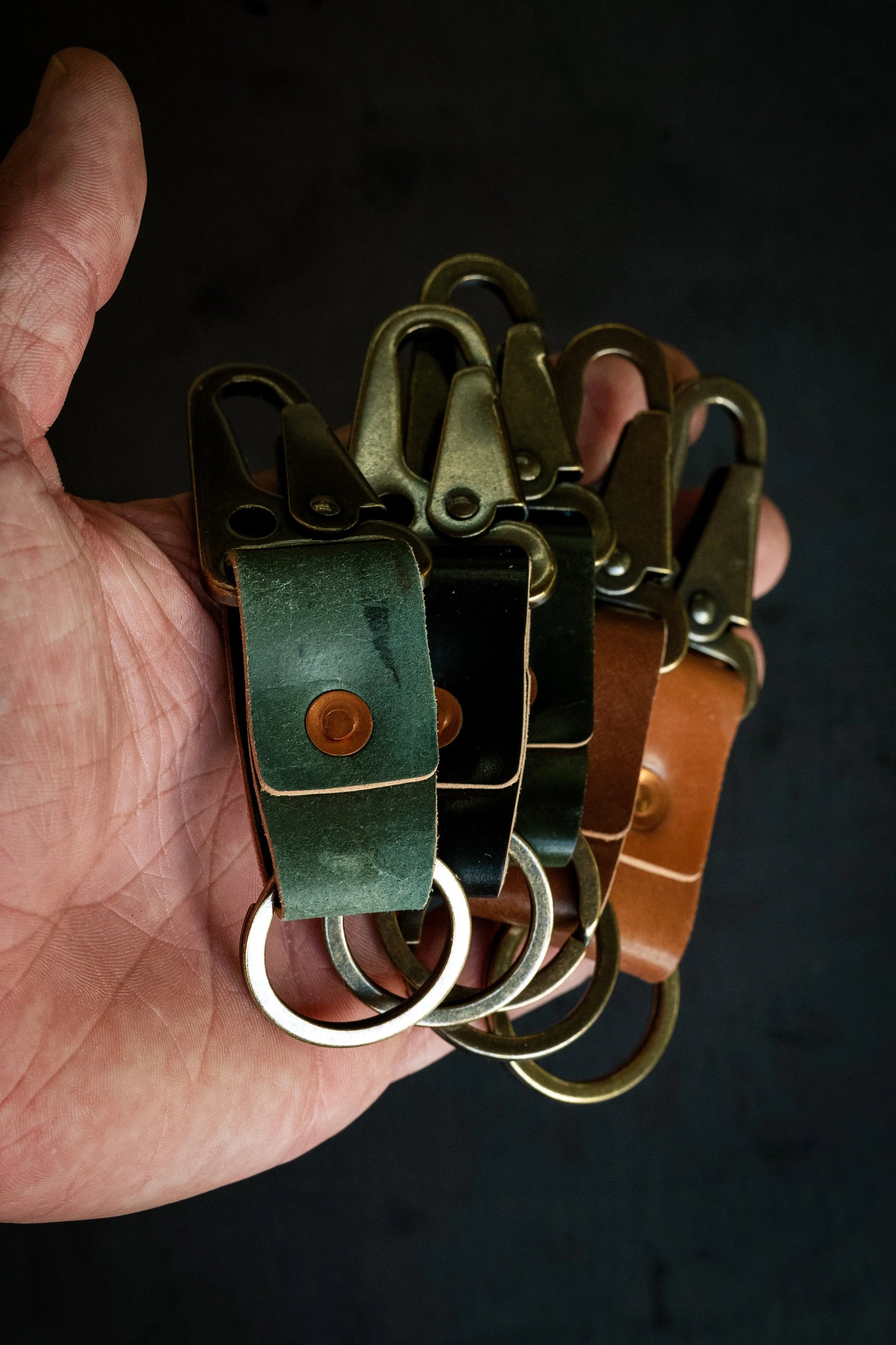 A hand holding four leather key clips with brass hardware, in green, black, brown, and tan leather.
