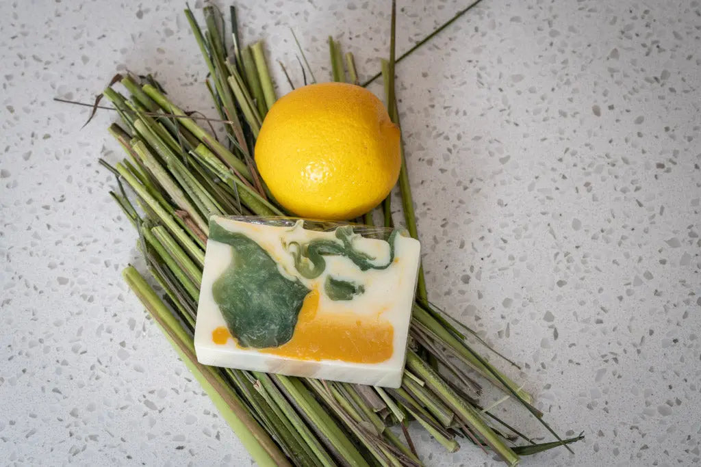 Bar of lemongrass goat milk soap slice with green and yellow swirls, next to a lemon and grass on a speckled surface.