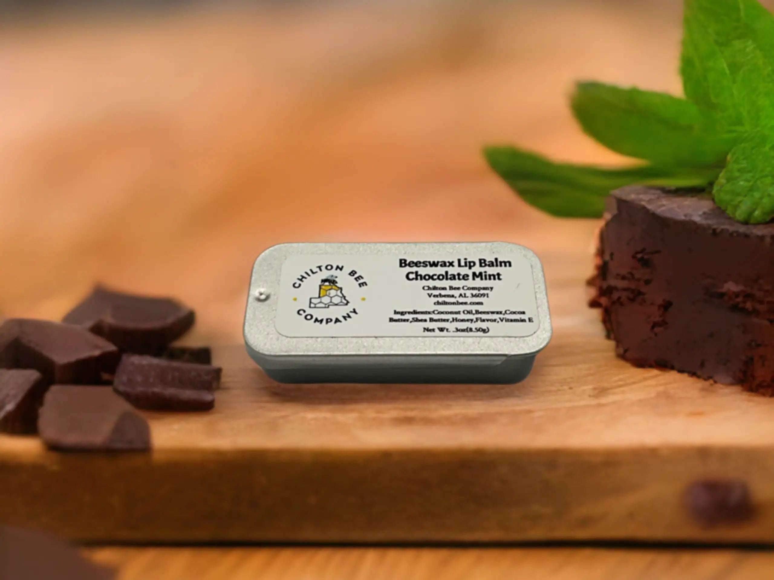Chilton Bee Company Chocolate Mint beeswax lip balm tin with chocolate and mint on a wood surface.