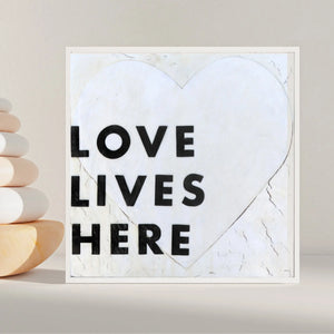 Framed art print with black "LOVE LIVES HERE" text on a white heart shape with textured background.