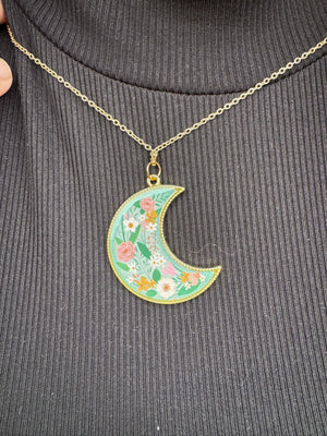 Floral moon pendant necklace on black ribbed fabric. Gold chain and trim, with pastel flower design inside a teal crescent moon shape.