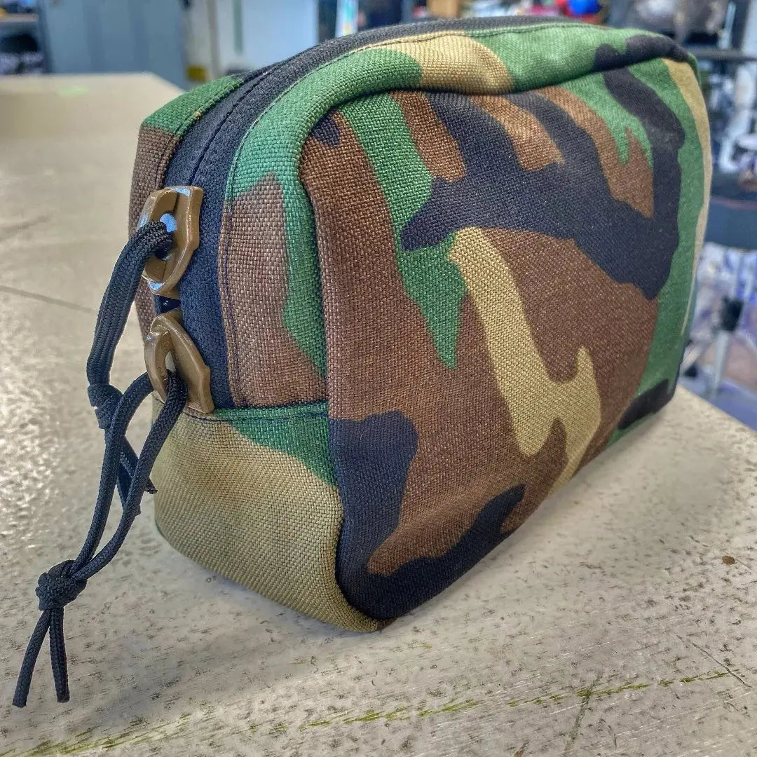 Woodland camouflage fanny pack with black cord zipper pulls and tan bottom.