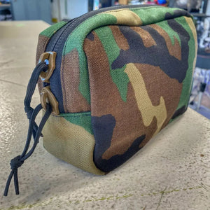 Woodland camouflage fanny pack with black cord zipper pulls and tan bottom.