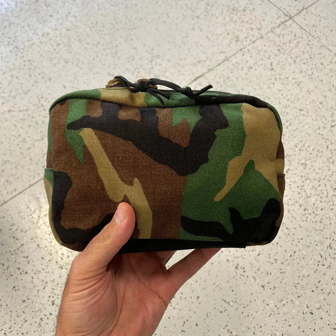 Hand holding a woodland camouflage fanny pack with a black zipper and drawstring.