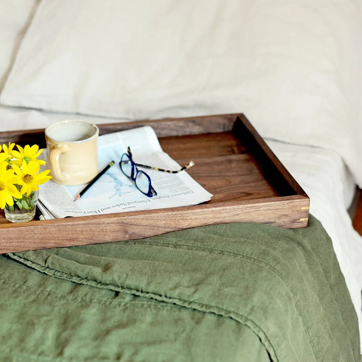 Wooden tray with brass corners holds a mug, newspaper, glasses, pencil, and yellow flowers on a green blanket and white bedding.