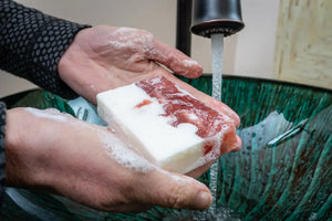 Hands washing a white and red marbled soap bar under running water.