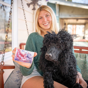 Woman holding a bar of pink and purple marbled goat's milk soap next to a black poodle.