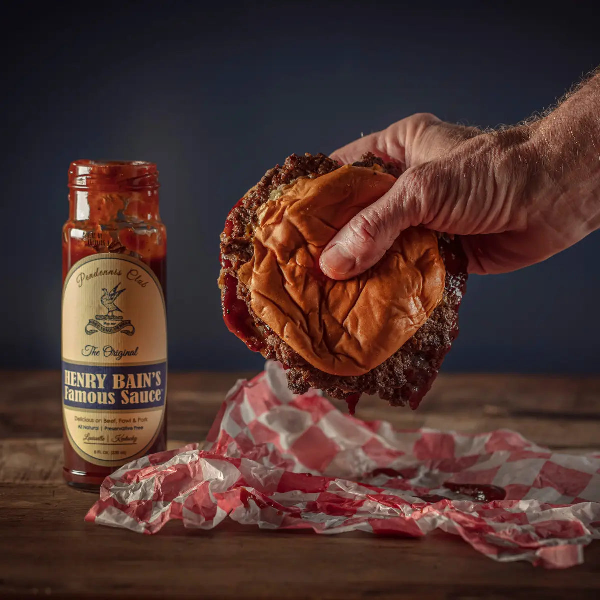 Hand holding a burger next to a bottle of Henry Bain's Famous Sauce on a wooden table.